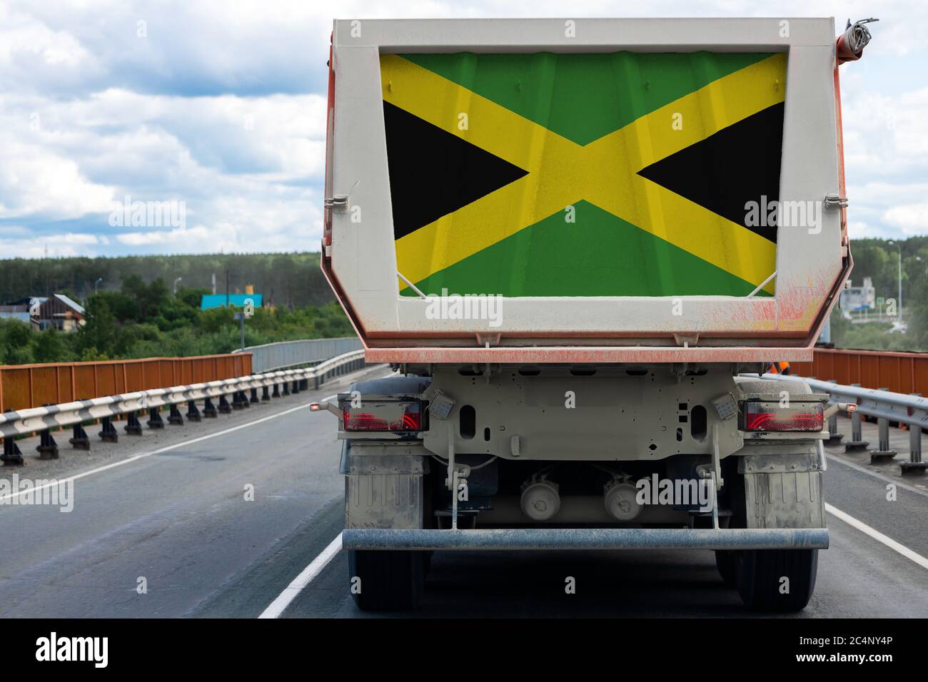 Big dirty truck with the national flag of Jamaica moving on the highway, against the background of the village and forest landscape. Concept of export Stock Photo