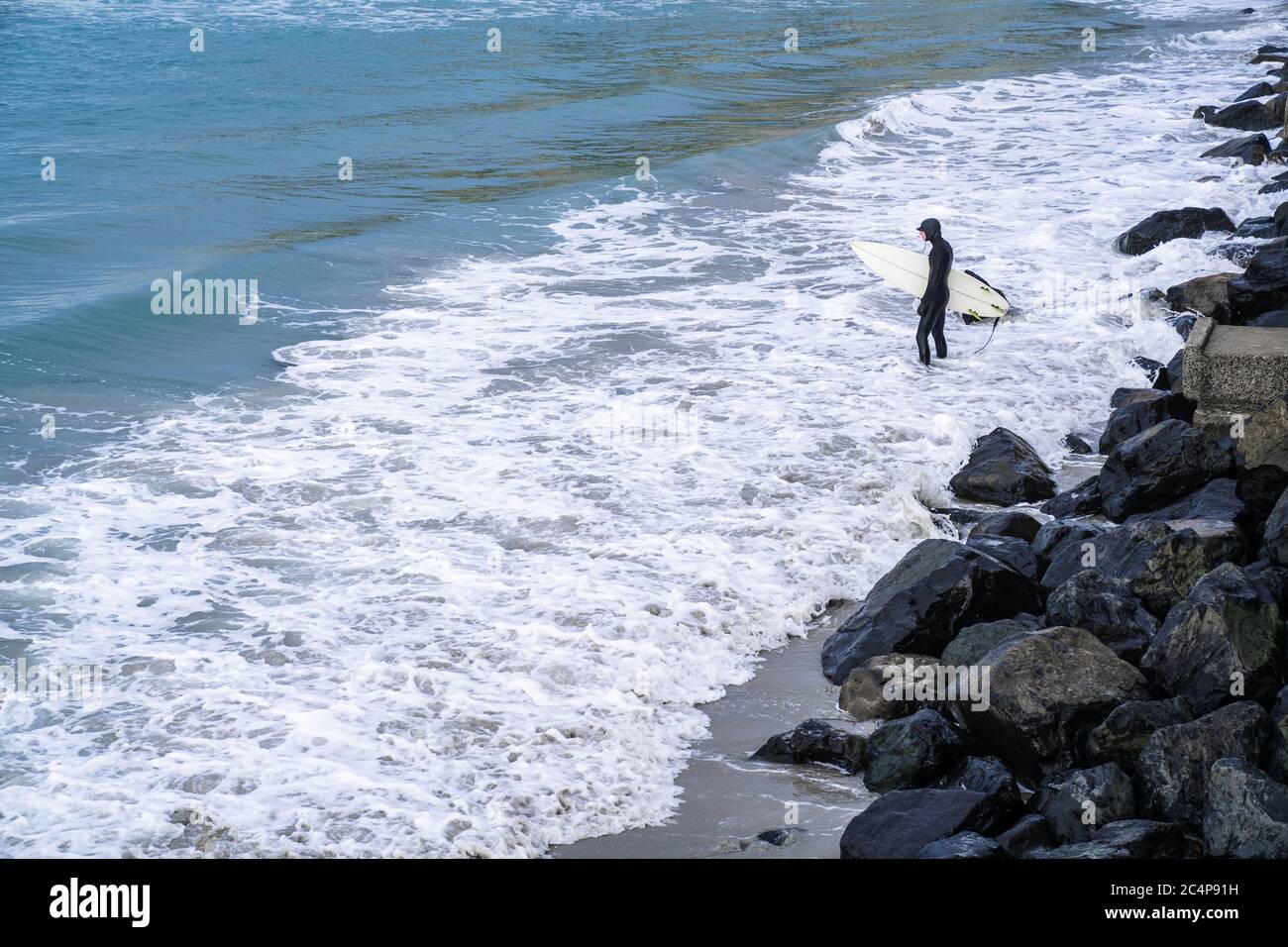 Surfer wearing wetsuit with surfboard watching ocean waves crash over rocks at St. Clair beach, Dunedin, New Zealand. Stock Photo