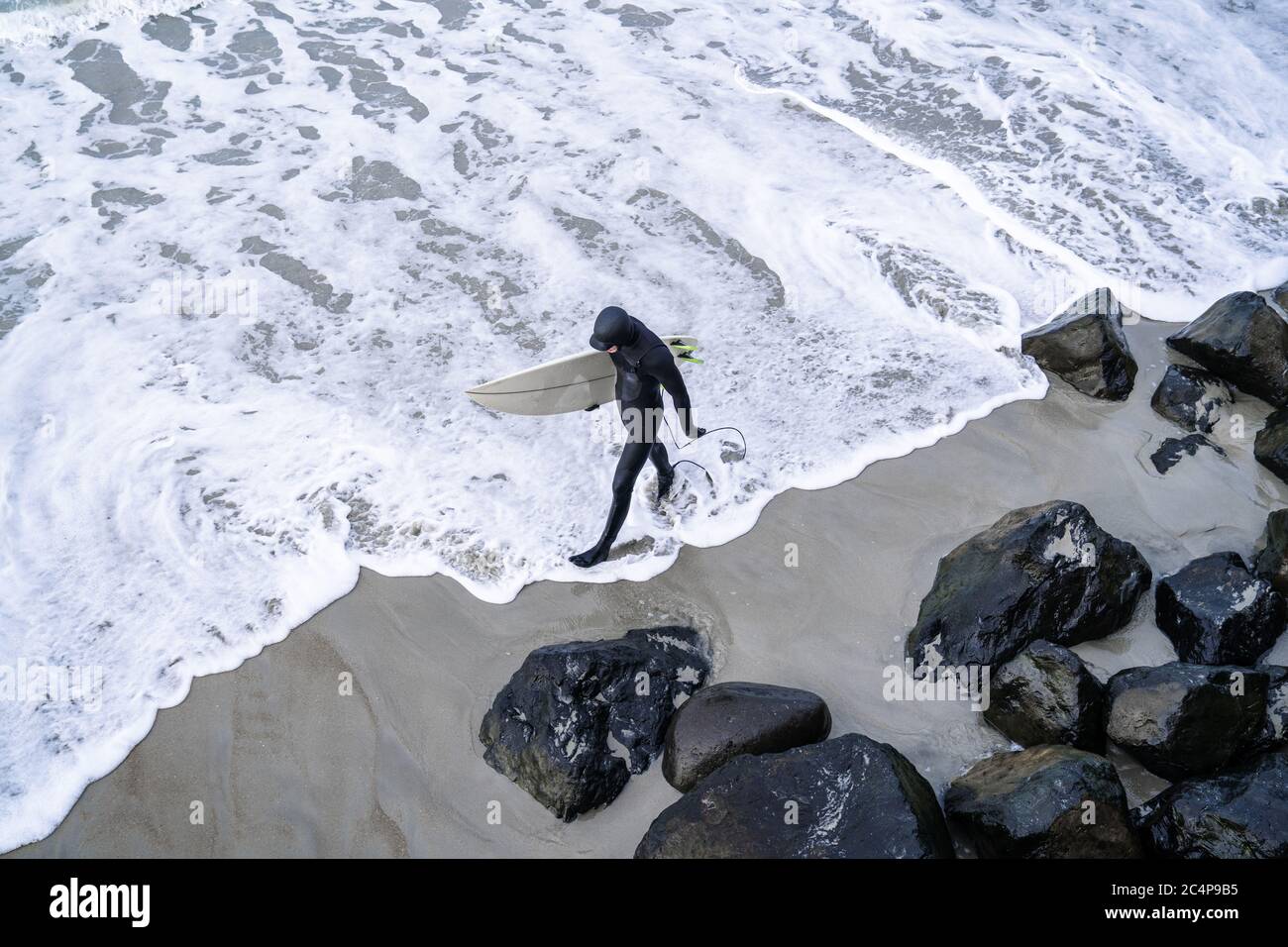 Surfer wearing wetsuit with surfboard watching ocean waves crash over rocks at St. Clair beach, Dunedin, New Zealand. Stock Photo