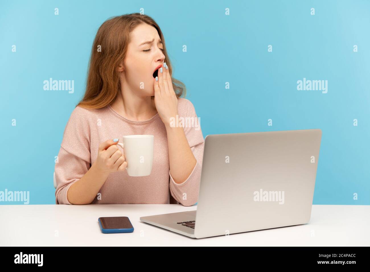 Exhausted drowsy woman sitting at workplace with laptop and yawning feeling sleepy, holding cup coffee to wake up, lazy inefficient employee at work. Stock Photo