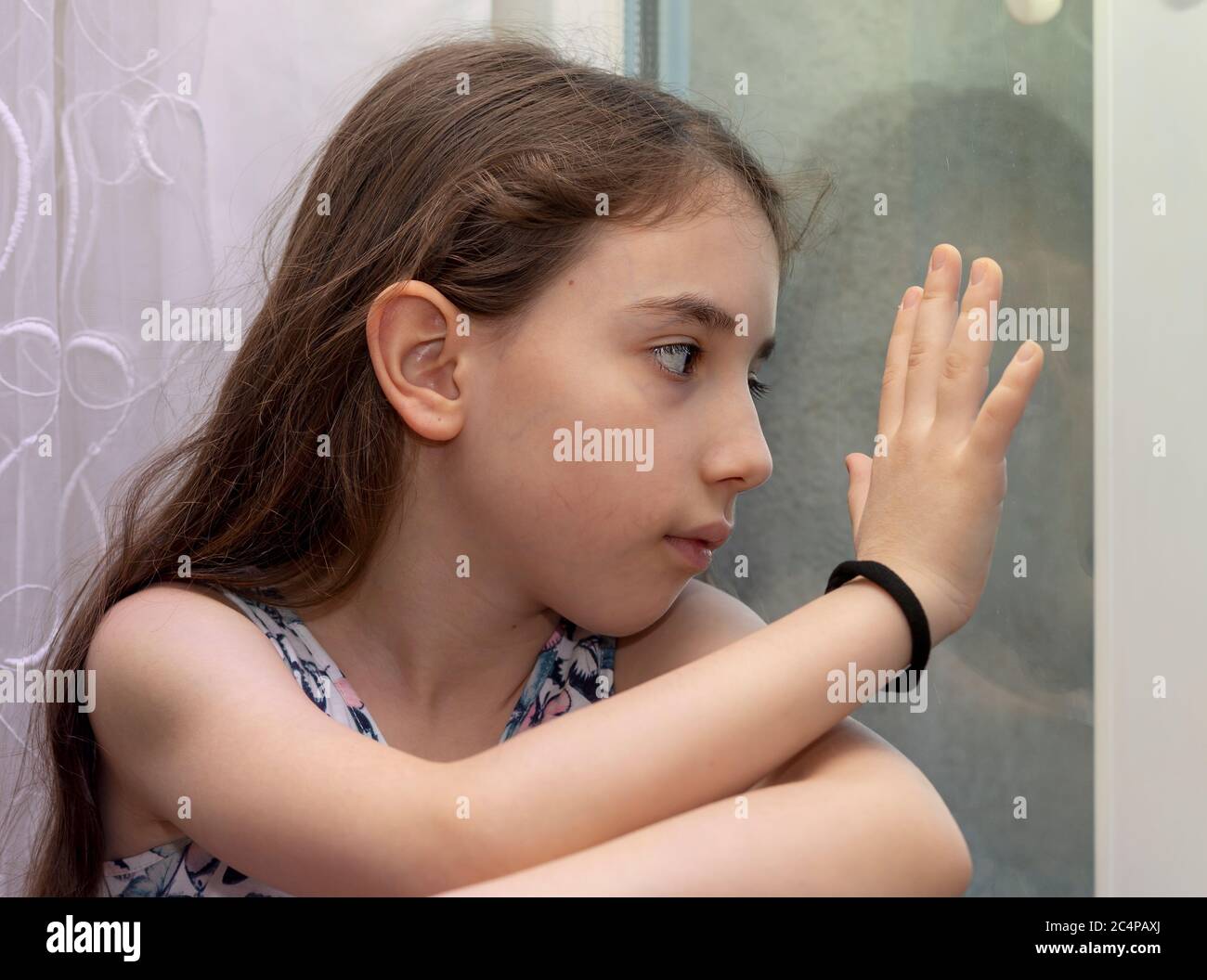 Unhappy and bored little girl waiting for behind the window.  A bored and depressed little girl put her head on the windows during corona quarantine Stock Photo