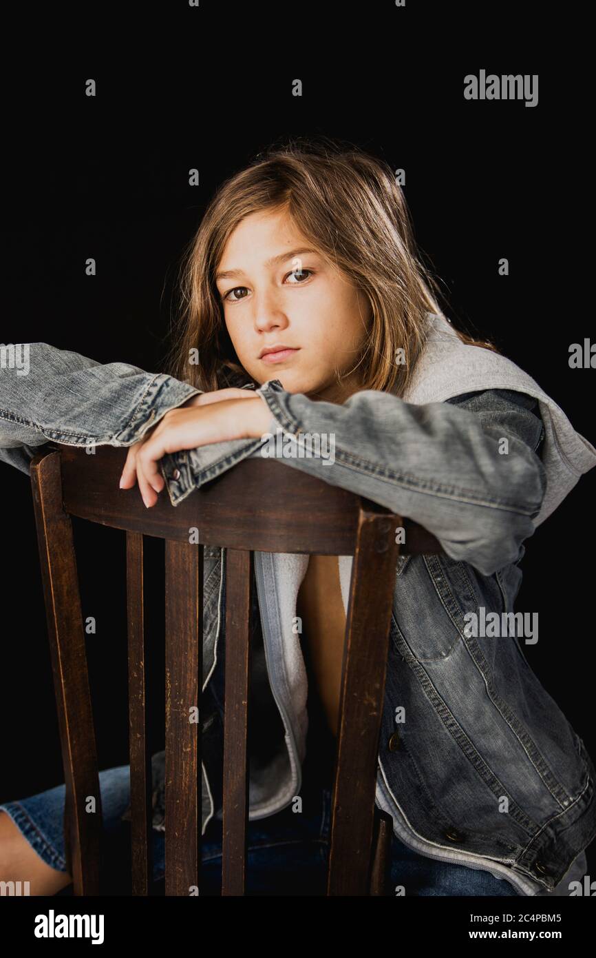 Eleven years old boy with hooded sweater and a jean jacket sitting on a wood chair against a black background Stock Photo