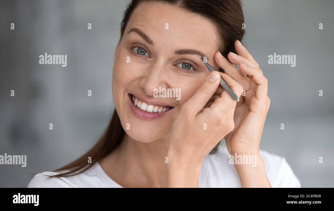 Young woman look in mirror pinch eyebrows with tweezers Stock Photo