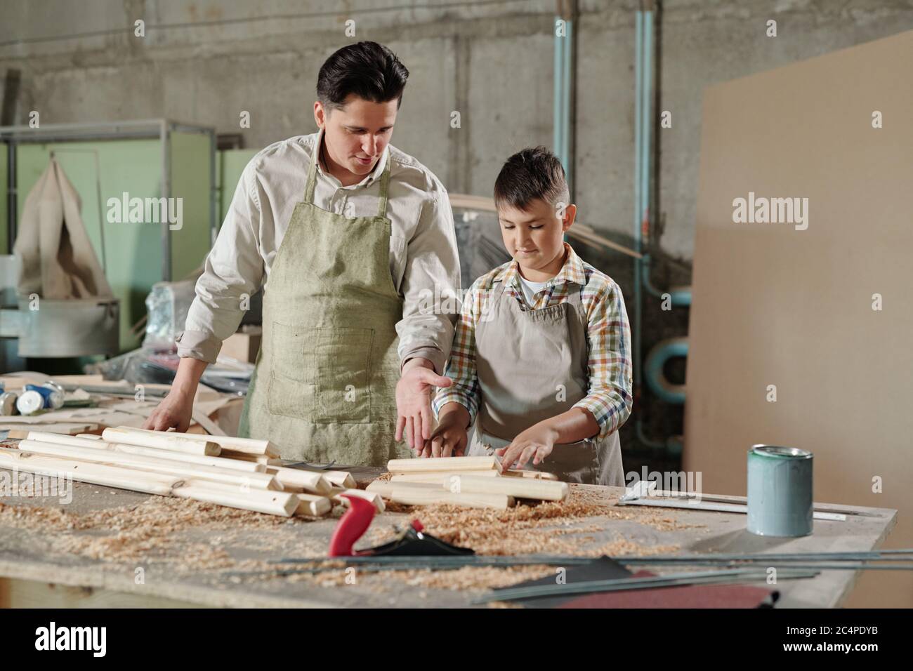 Male carpenter in apron pointing at planks while explaining teenage son ...