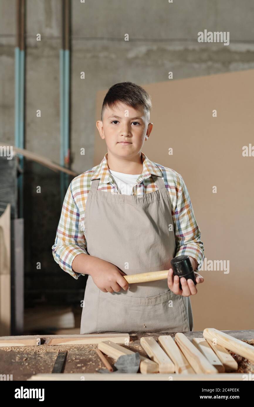 Portrait of serious teenage boy in apron standing with hammer at desk ...