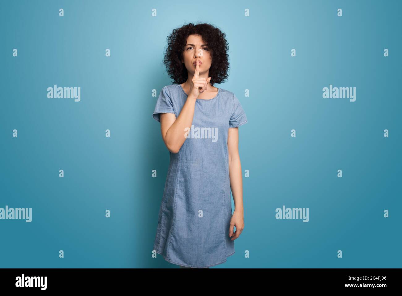 Monochrome photo of a caucasian woman with curly hair gesturing the silence sign on a blue studio wall Stock Photo