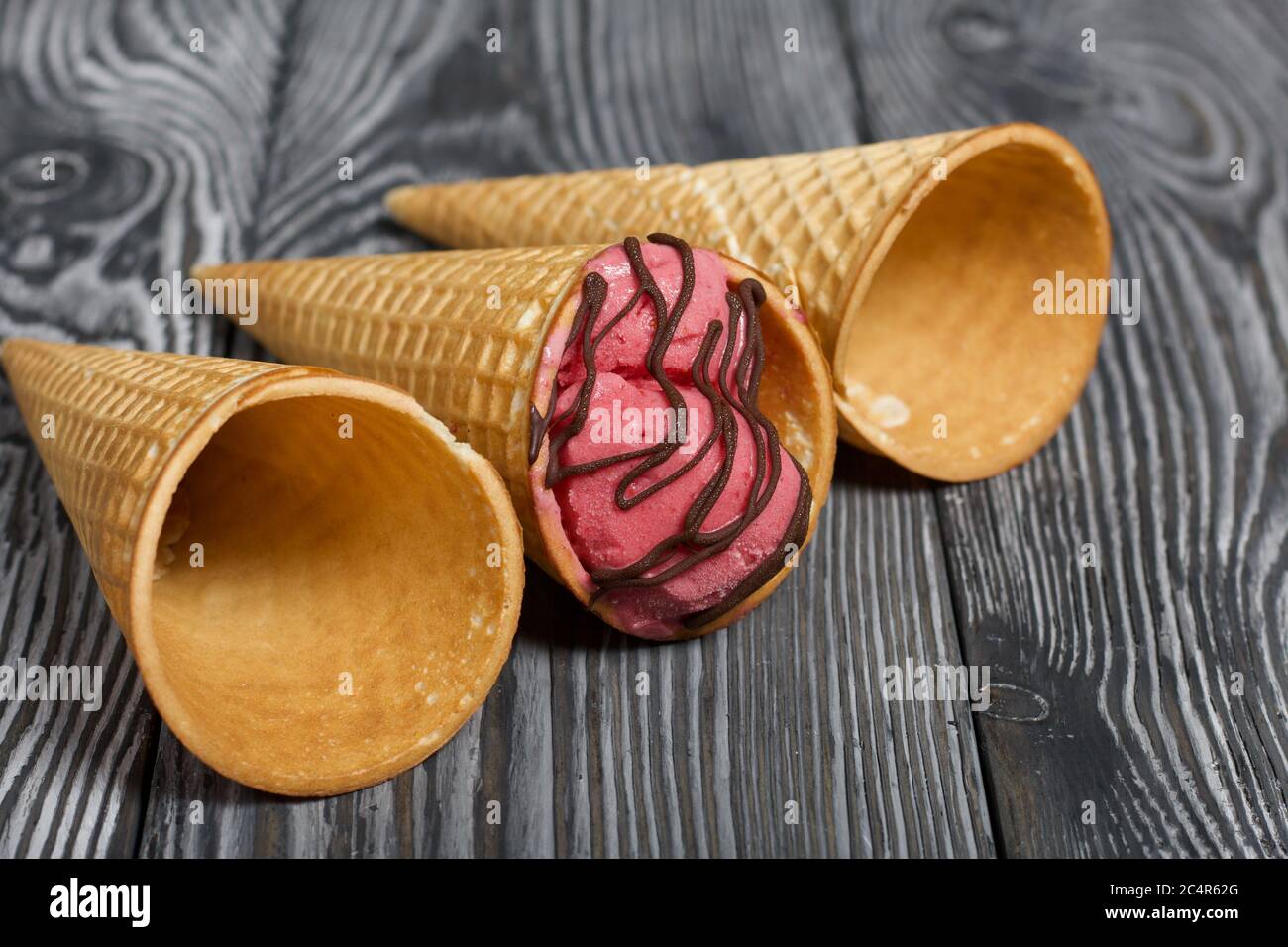 Strawberry ice cream in a waffle cone. Garnished with Chocolate. Nearby are empty waffle cones without ice cream. On pine boards painted in black and Stock Photo