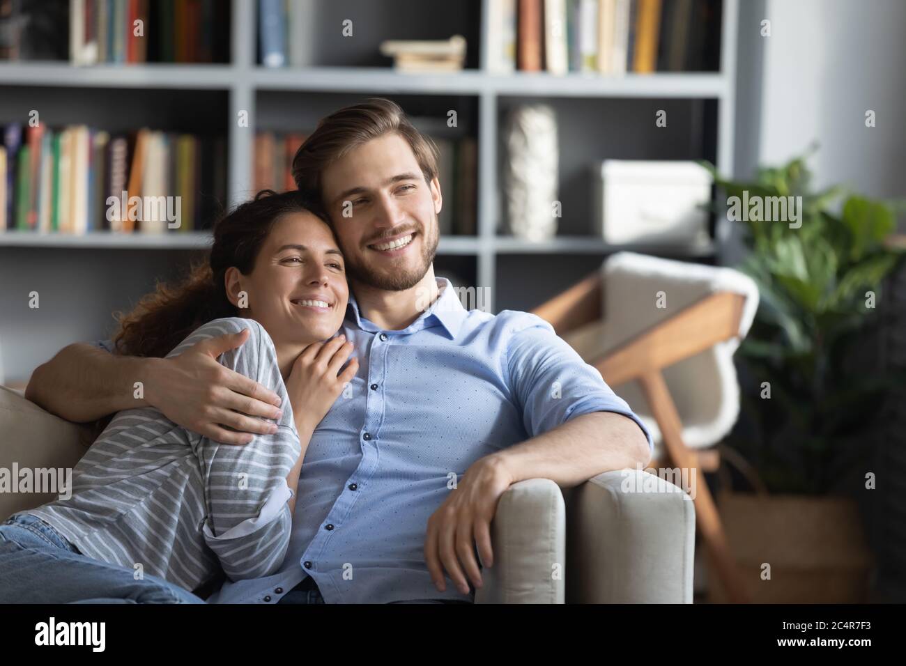 Smiling dreamy young couple hugging, relaxing on couch at home Stock