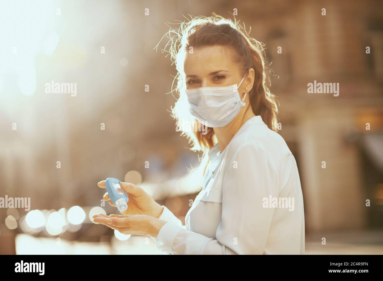 Life during coronavirus pandemic. modern 40 years old woman in blue blouse with medical mask disinfecting hands with sanitizer outdoors in the city. Stock Photo