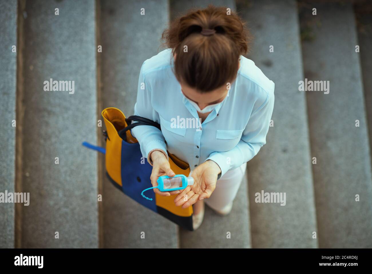 Life during coronavirus pandemic. Upper view of trendy woman in blue blouse with medical mask and handbag disinfecting hands with an antibacterial age Stock Photo