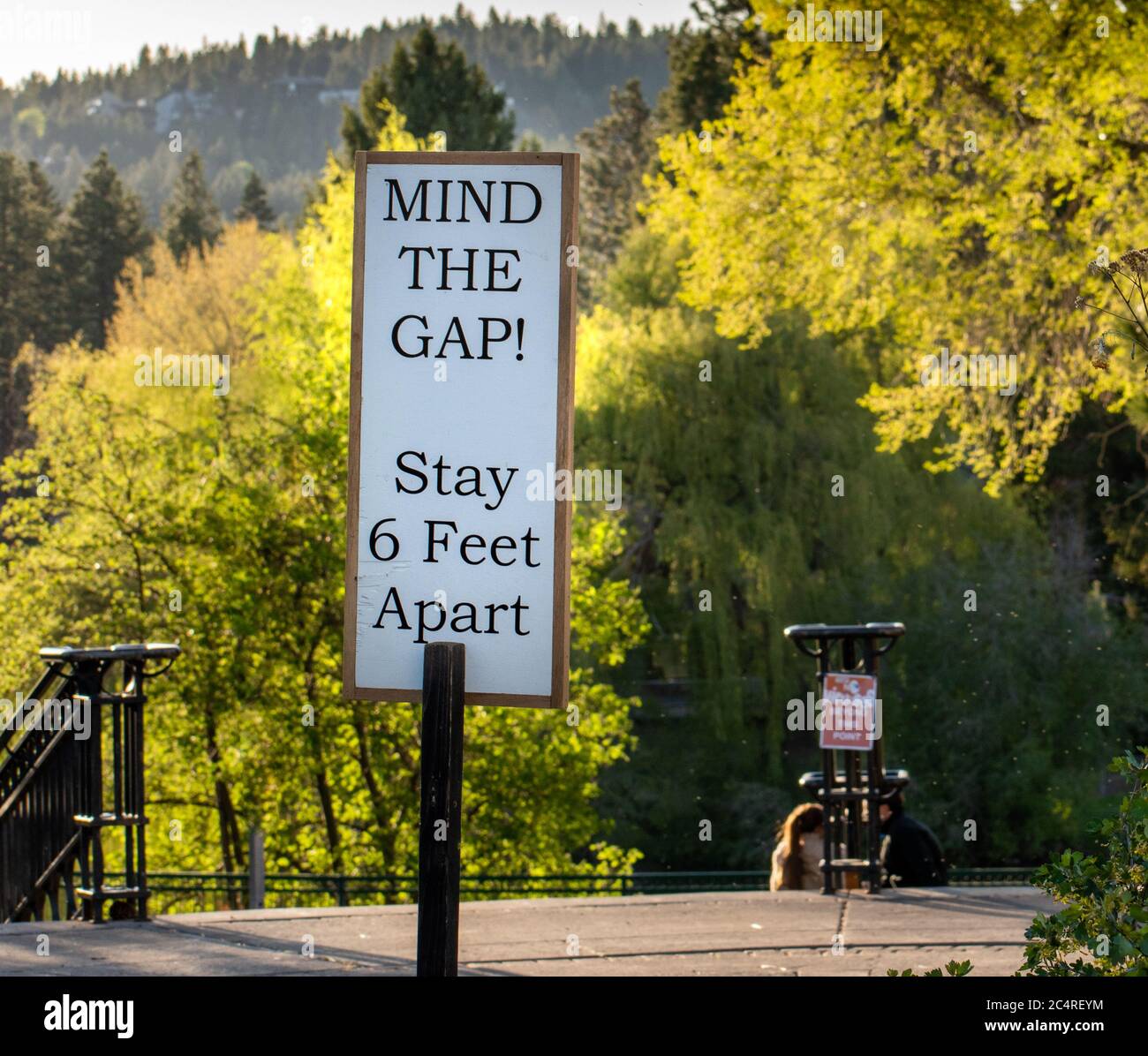 Social distancing sign reads Mind The Gap at Bend Oregon park entrance while two people sit closely next to each other. Stock Photo