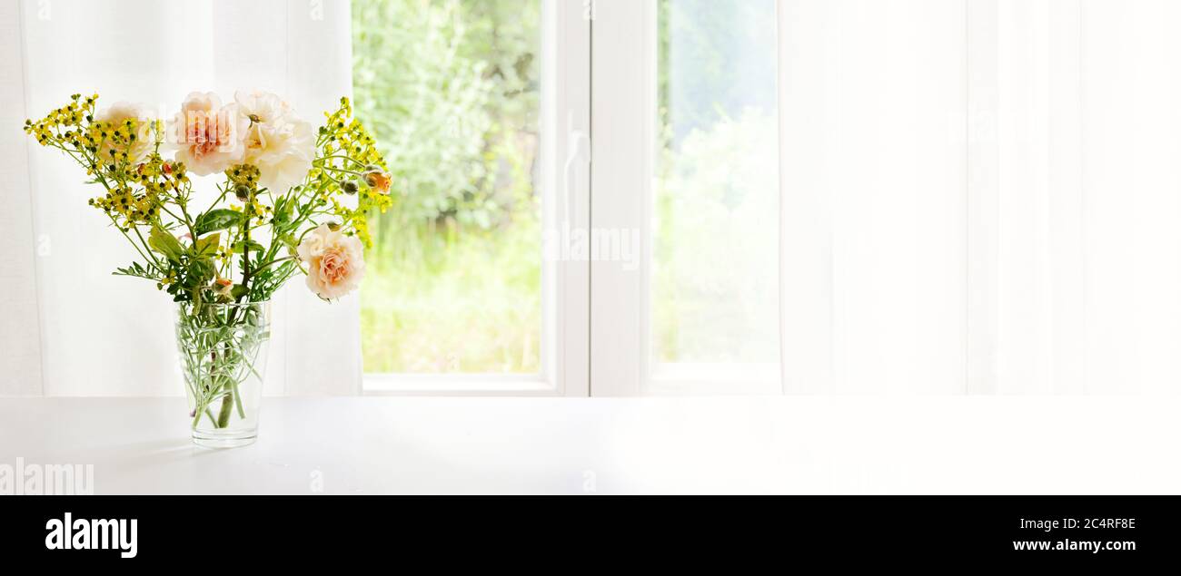 Bouquet from orange roses and rue in glass vase at the window in front of a white curtain, panoramic format, large copy space, selected focus Stock Photo