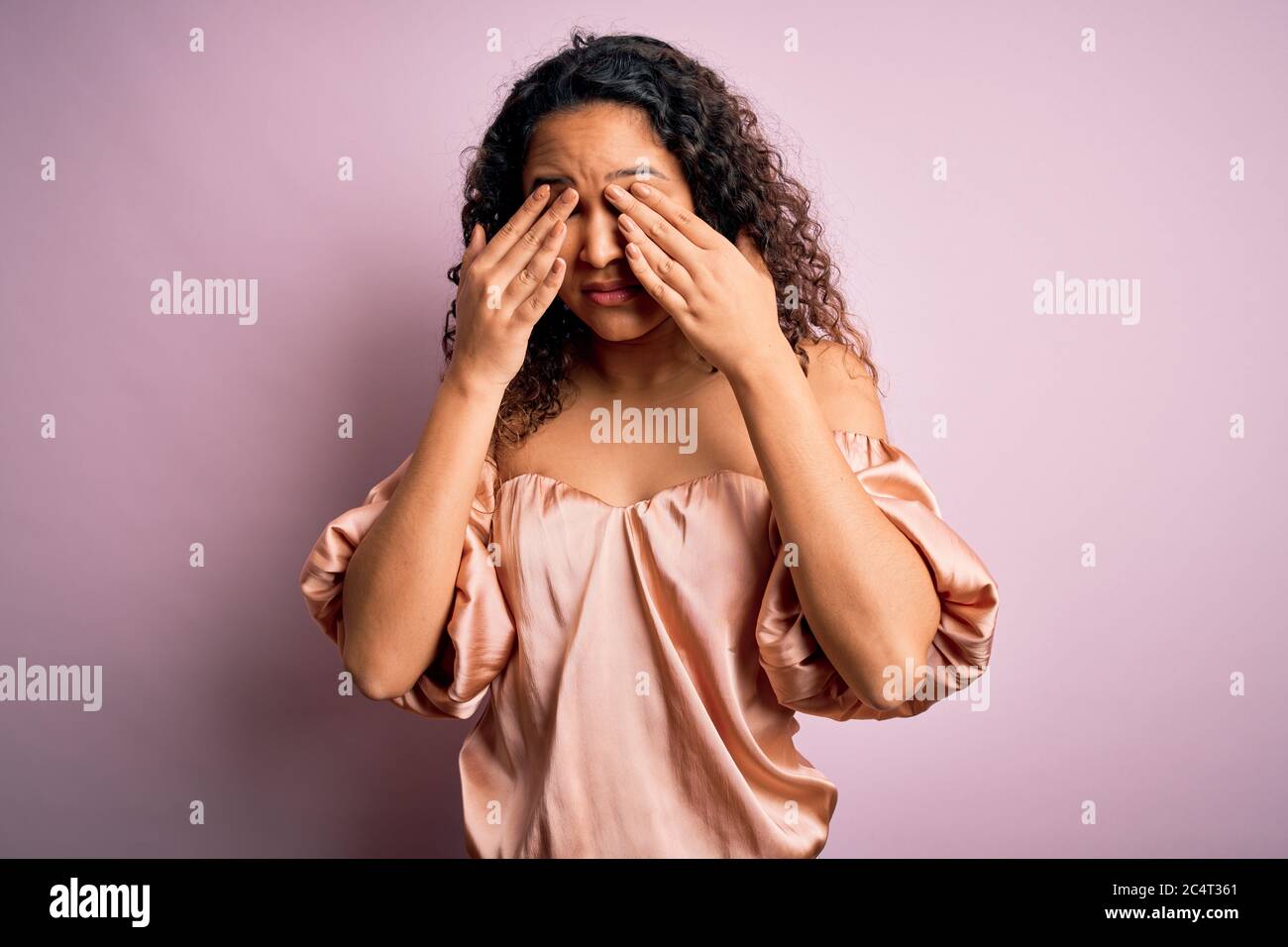 Young beautiful woman with curly hair wearing casual t-shirt standing over pink background rubbing eyes for fatigue and headache, sleepy and tired exp Stock Photo