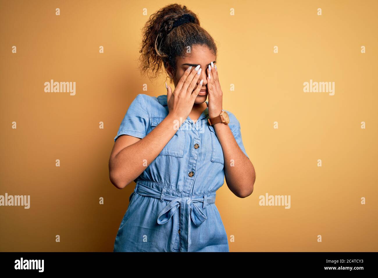 Young beautiful african american girl wearing denim dress standing over yellow background rubbing eyes for fatigue and headache, sleepy and tired expr Stock Photo
