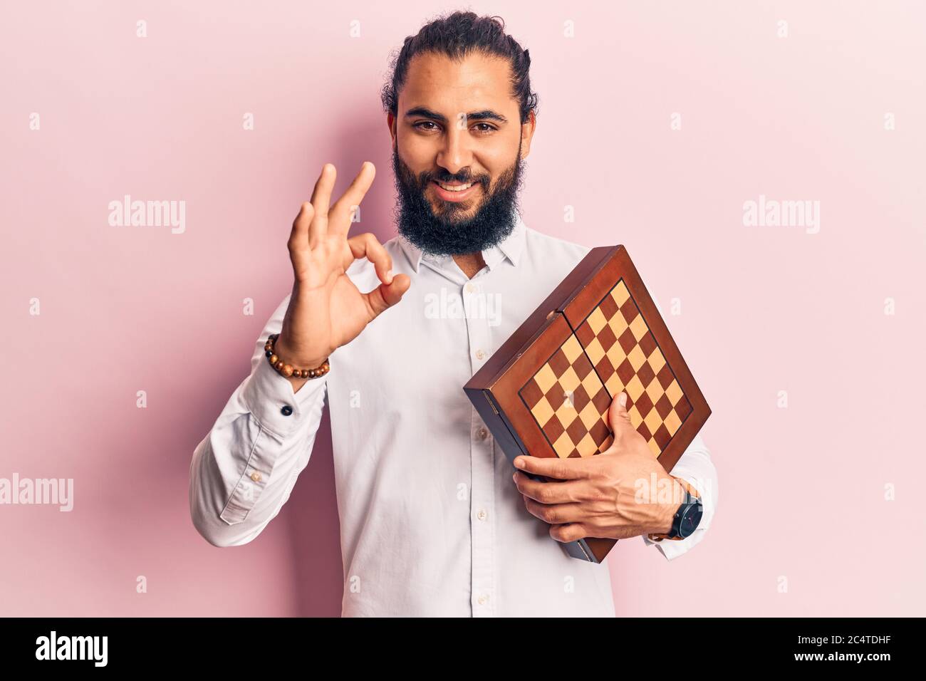 Young arab man holding chess doing ok sign with fingers, smiling friendly gesturing excellent symbol Stock Photo
