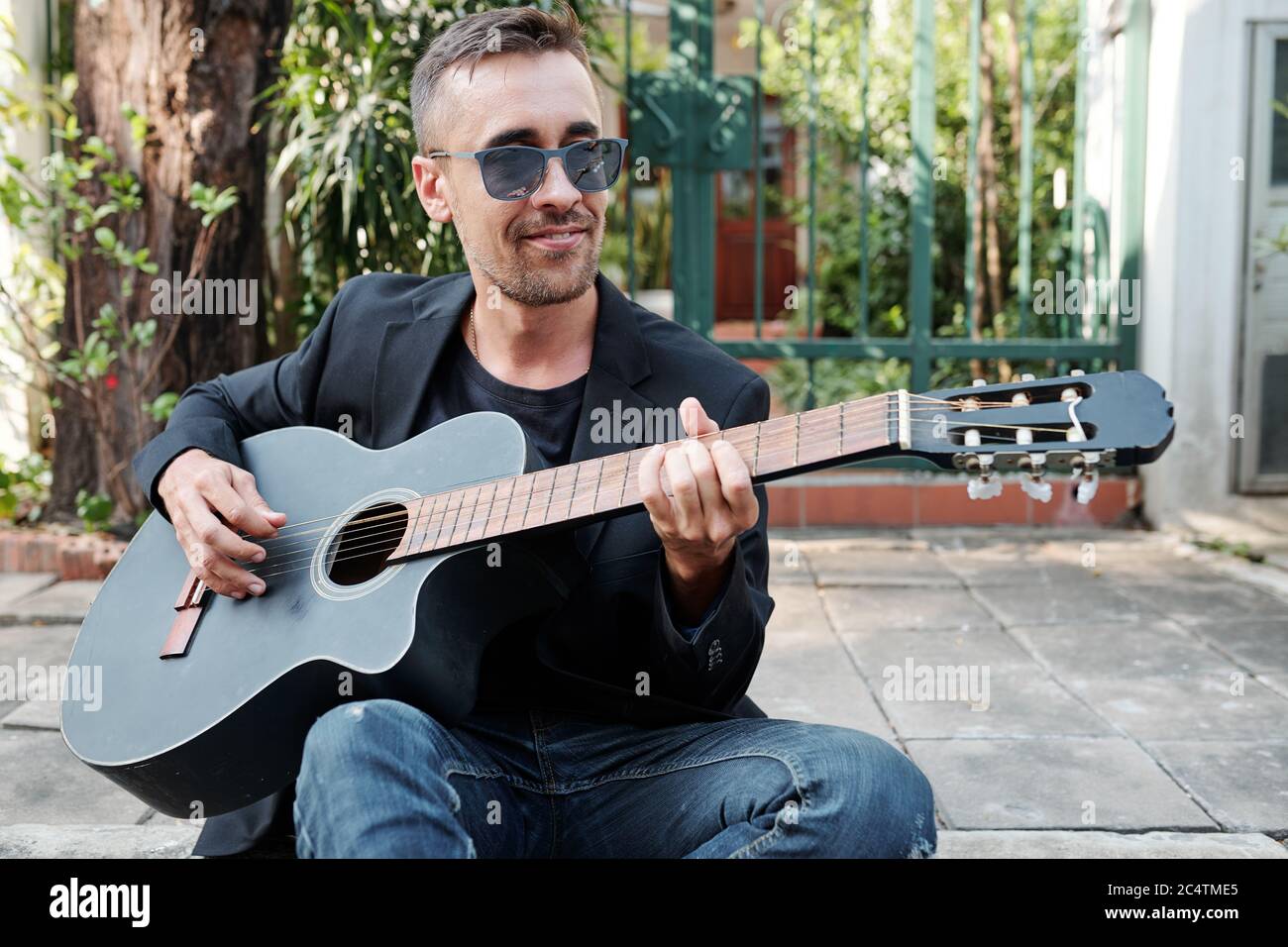 Smiling handsome man in sunglasses sitting on pavement and playing guitar Stock Photo