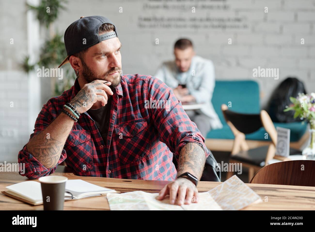 Pensive young bearded man sitting at table in cafe and looking away while thinking about new trip Stock Photo