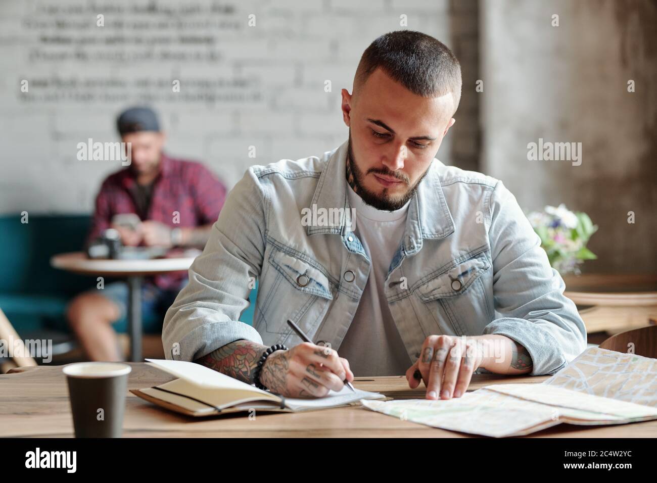 Concentrated hipster guy with beard sitting at table in modern cafe and viewing paper map while making notes about trip Stock Photo
