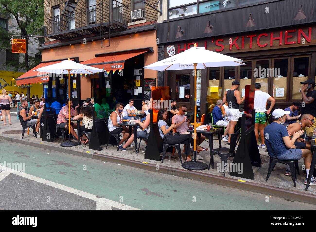 New York, NY. June 28, 2020. People holding drinks and conversation outside 'Ritas on Ninth Avenue in the Hells Kitchen neighborhood. Tables with colorful partitions have been setup on the sidewalk for socially distanced outdoor dining under Phase 2 reopening of New York City. Stock Photo
