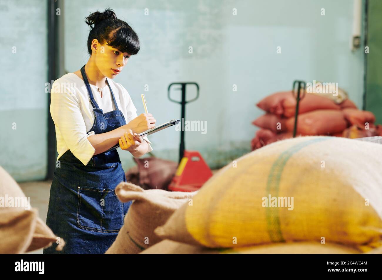 Young female warehouse worker checking sacks and taking notes in document on clipboard Stock Photo