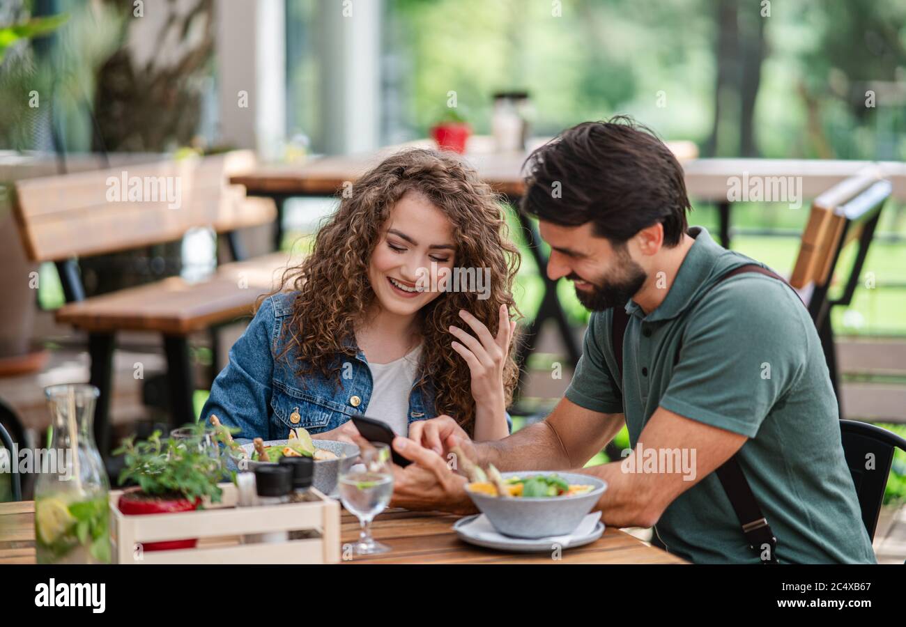 Happy couple sitting outdoors on terrace restaurant, using smartphone. Stock Photo