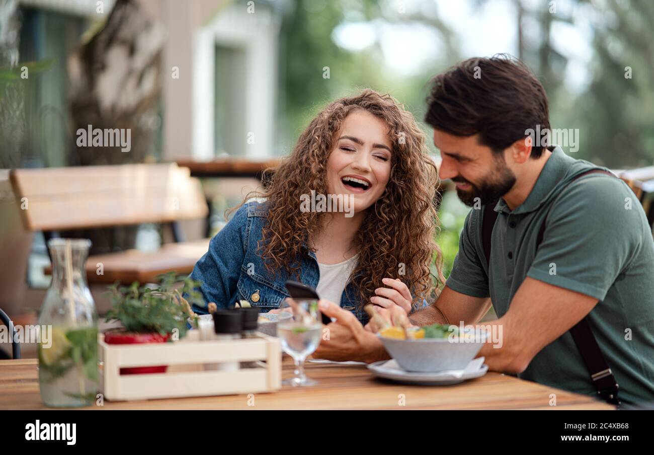 Happy couple sitting outdoors on terrace restaurant, using smartphone. Stock Photo