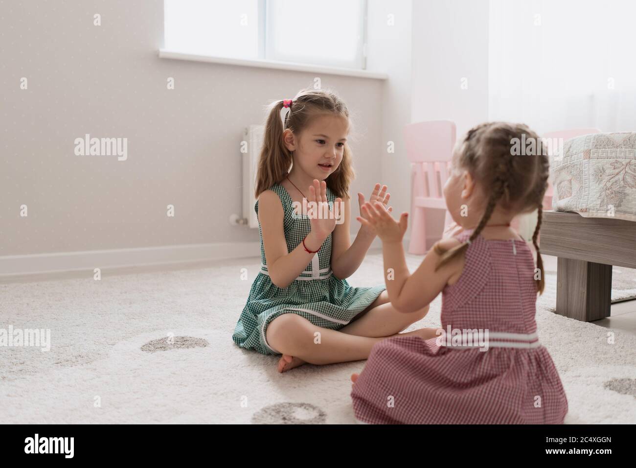 Cute little sisters playing together sitting on the carpet in identical dresses different colors in a room with lots of light Stock Photo
