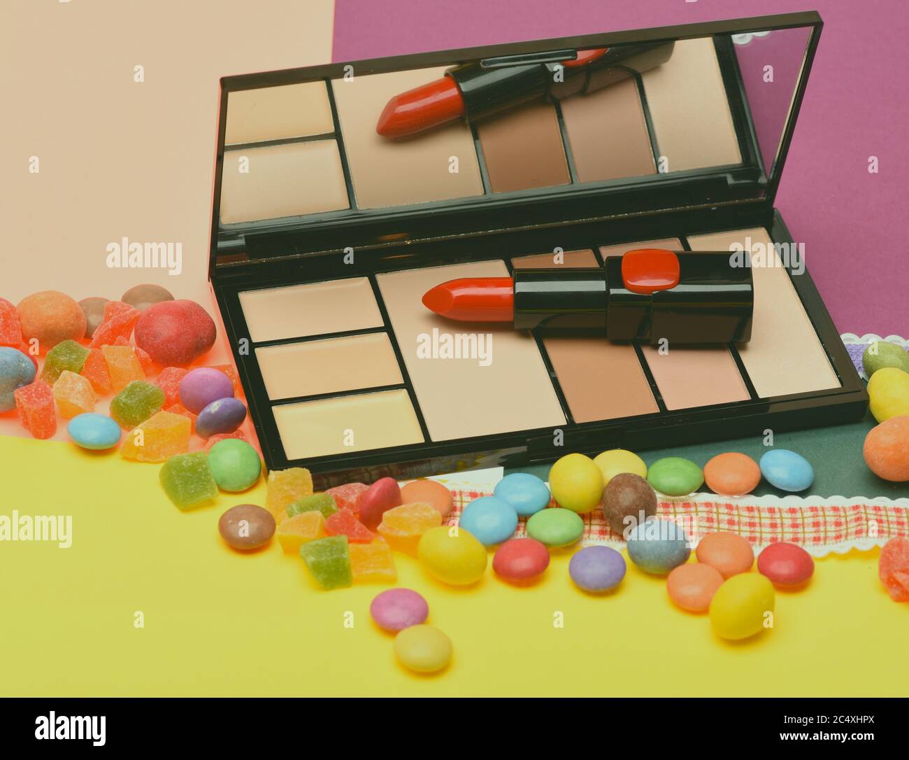 Red, scarlet lipstick and palette with mirror lay on peachy and yellow background near dragee sweets and candied fruit. Beauty supplies, lipstick, palette, powder, rouge, concealer. Make up concept. Stock Photo