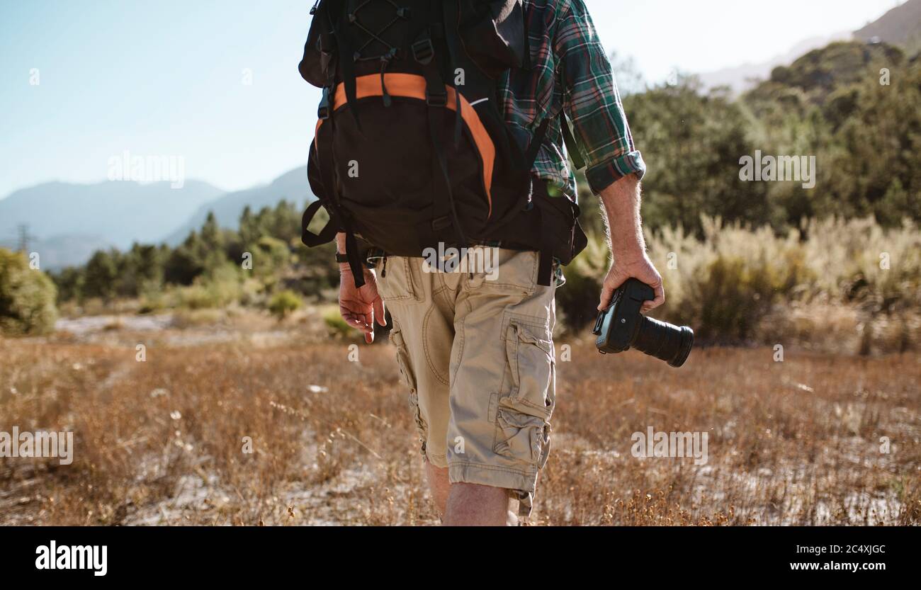 Rear view of a senior man carrying a backpack hiking in nature holding a digital camera. Man on hiking trip. Stock Photo