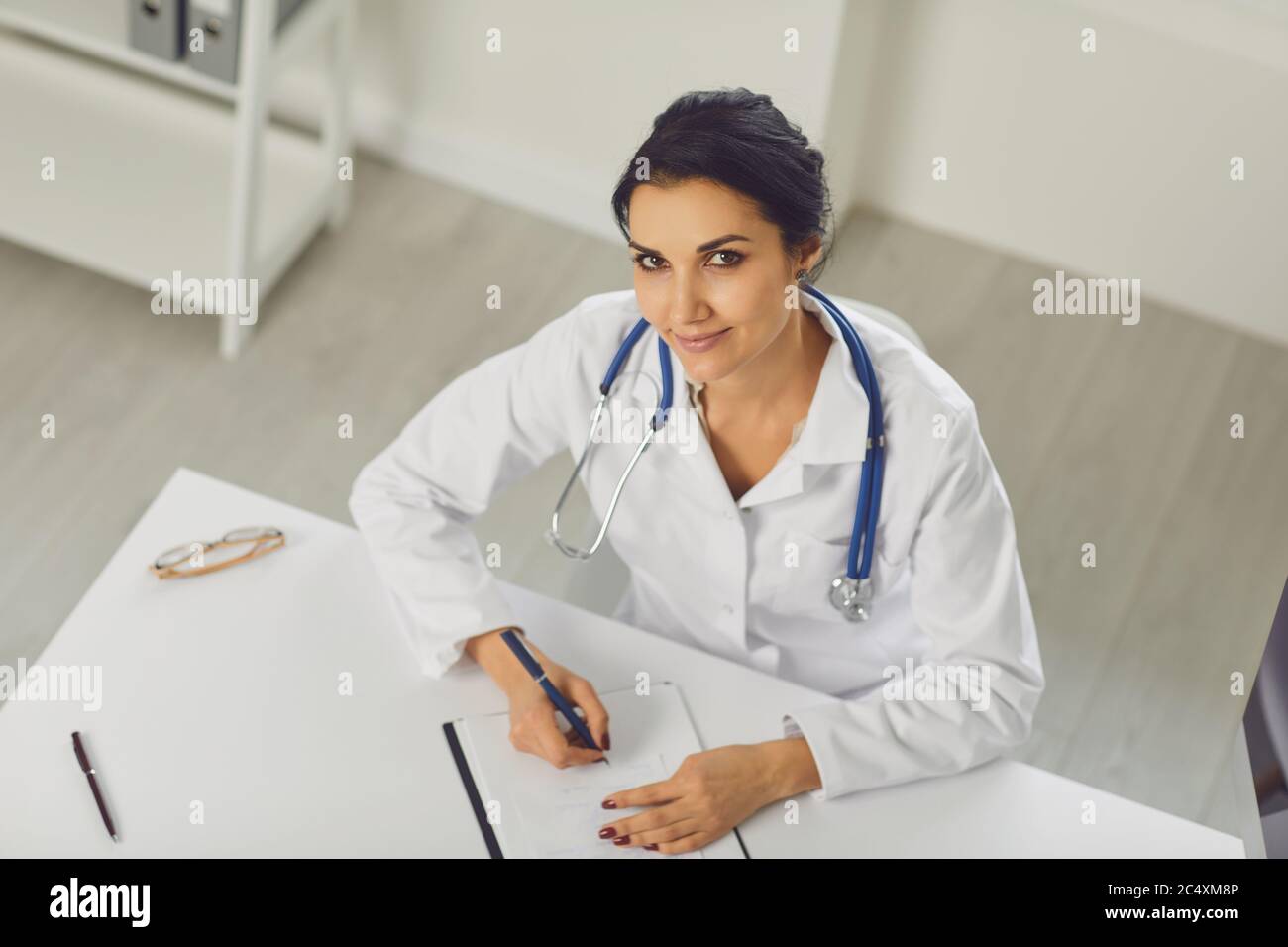 Confident woman doctor pediatrician writes in a clipboard sitting at a table in a white office of the hospital Stock Photo