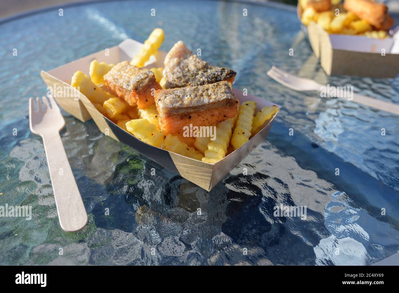 fish and chips with fried smoked salmon and french fries in a cardboard box and a wooden fork on a glass table, fast food in an outdoor street restaur Stock Photo