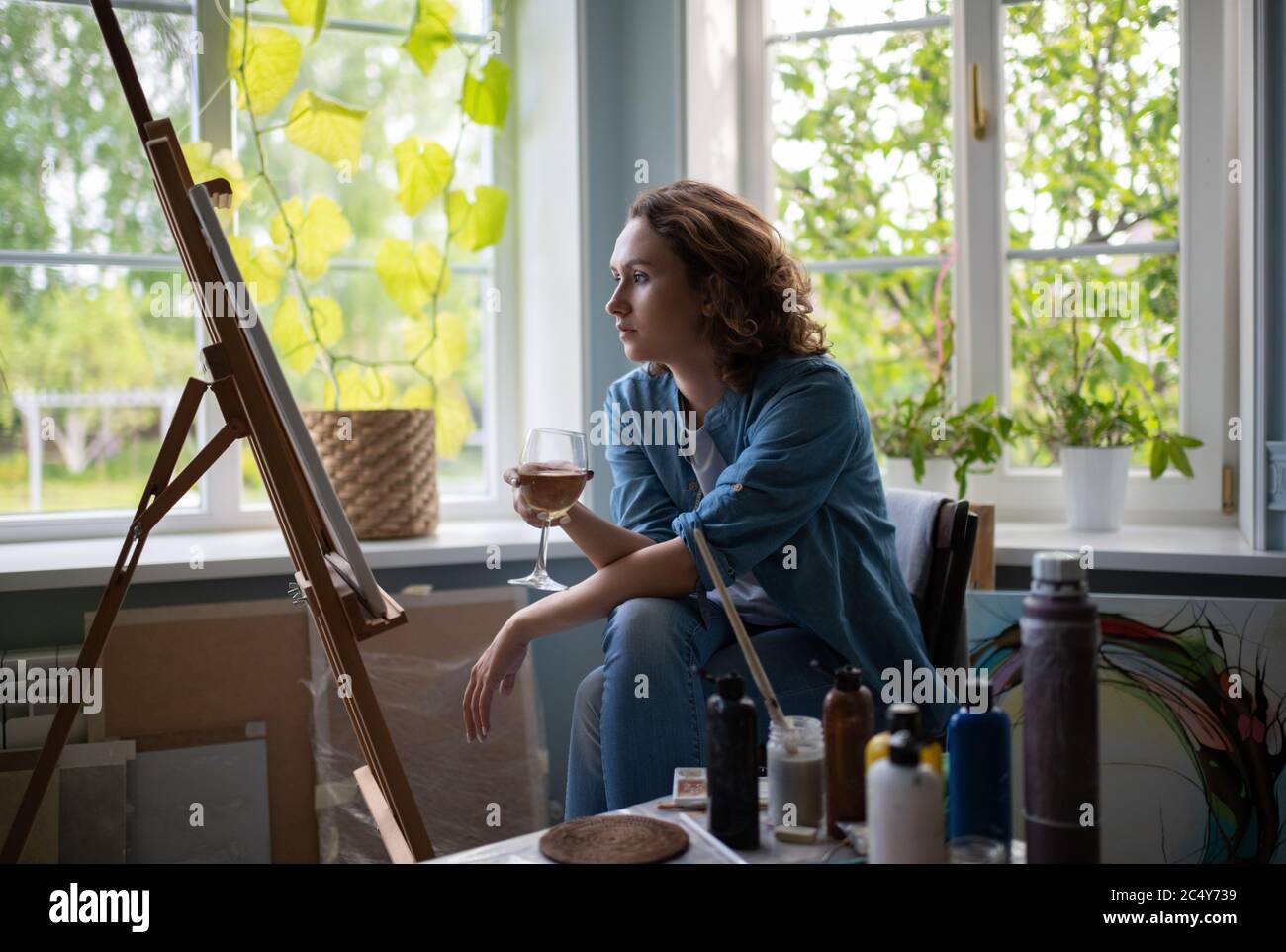 Adult artist with wine admiring artwork on easel against window in cozy studio Stock Photo