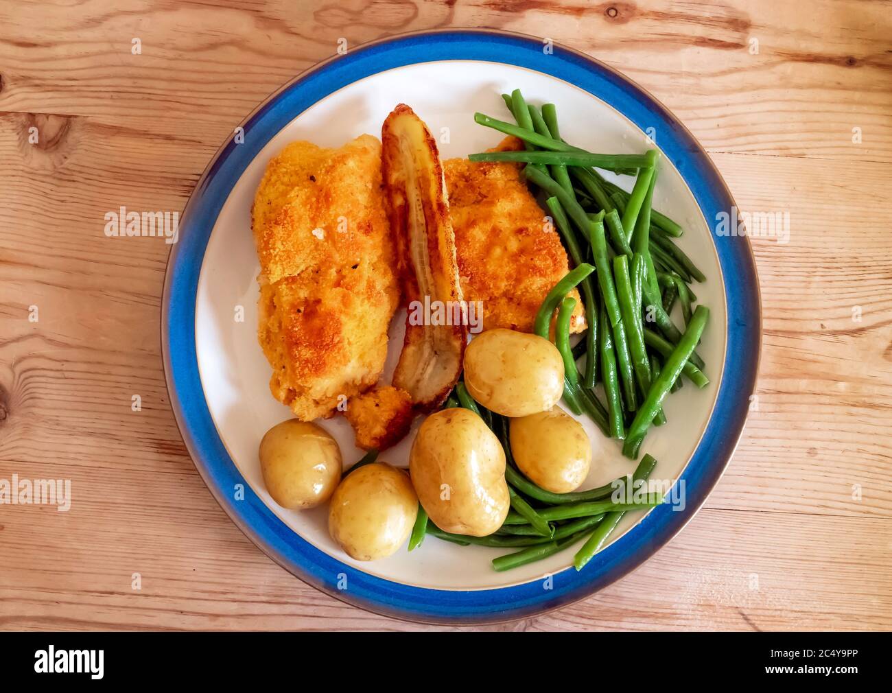 Lunch main course breaded chicken fillets with green beans new potatoes  and a fried bannana white plate blue edge wooden table top Stock Photo