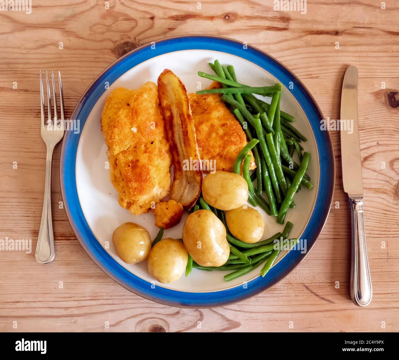 Lunch main course breaded chicken fillets with green beans new potatoes  and a fried banana white plate blue edge wooden table top with cutlery Stock Photo