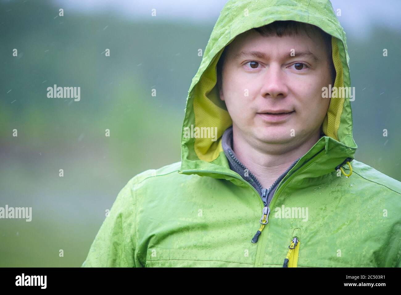 A young guy in a raincoat with a hood stands in the rain. Raindrops