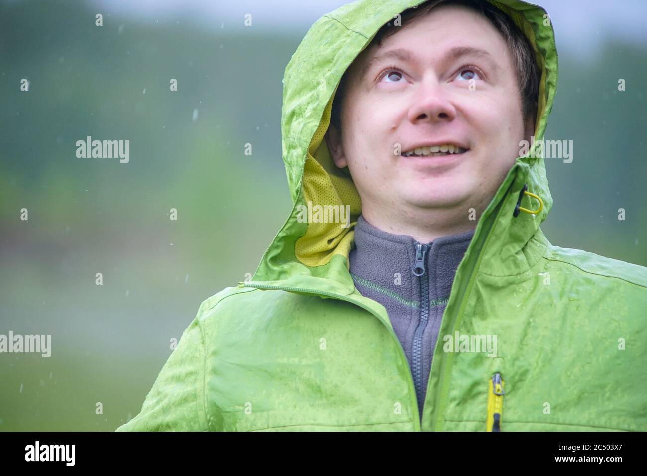 A young guy in a raincoat with a hood stands in the rain. Raindrops fall on your face and jacket. Stock Photo