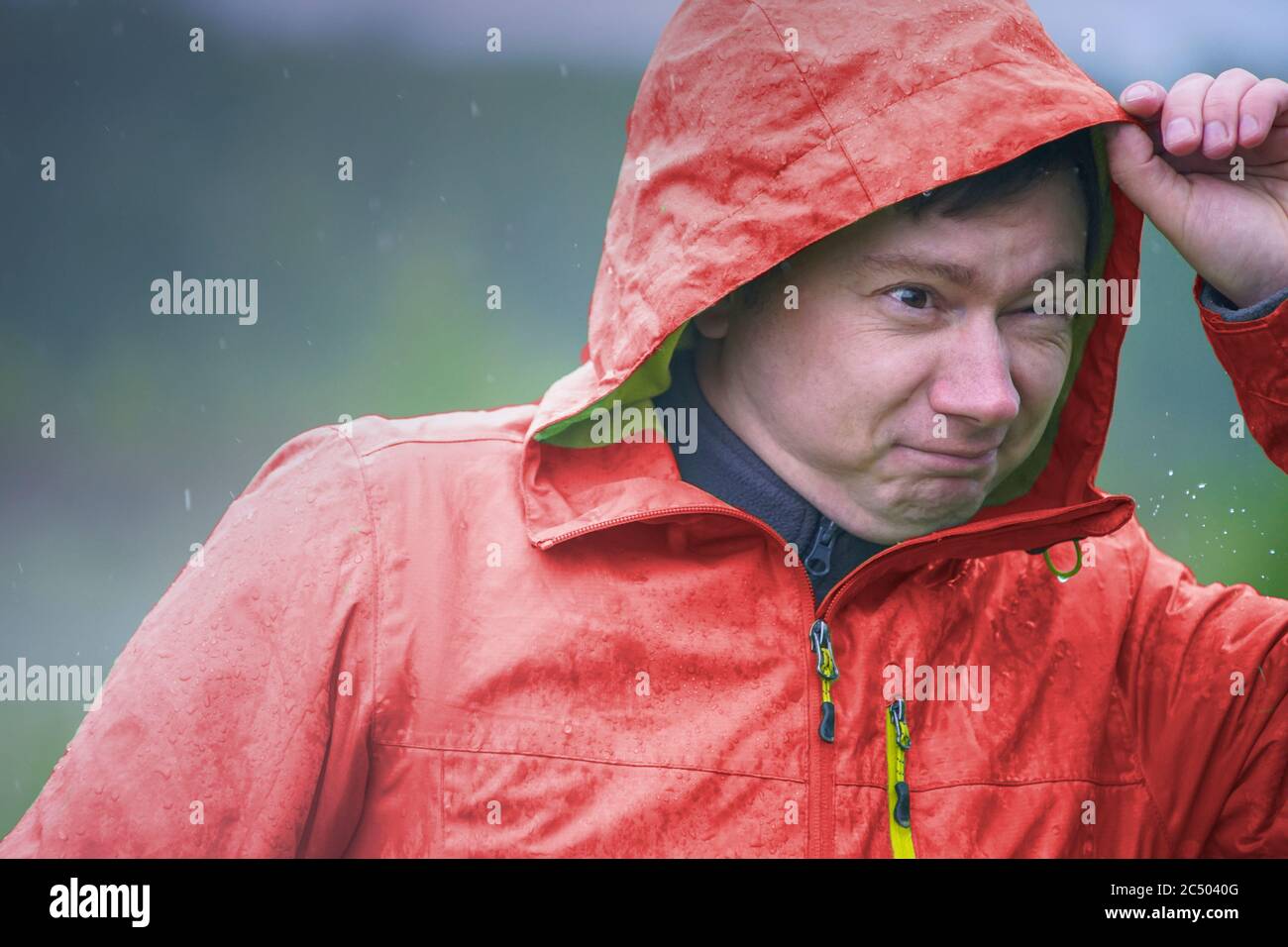 A young guy in a raincoat with a hood stands in the rain. Raindrops fall on your face and jacket. Stock Photo