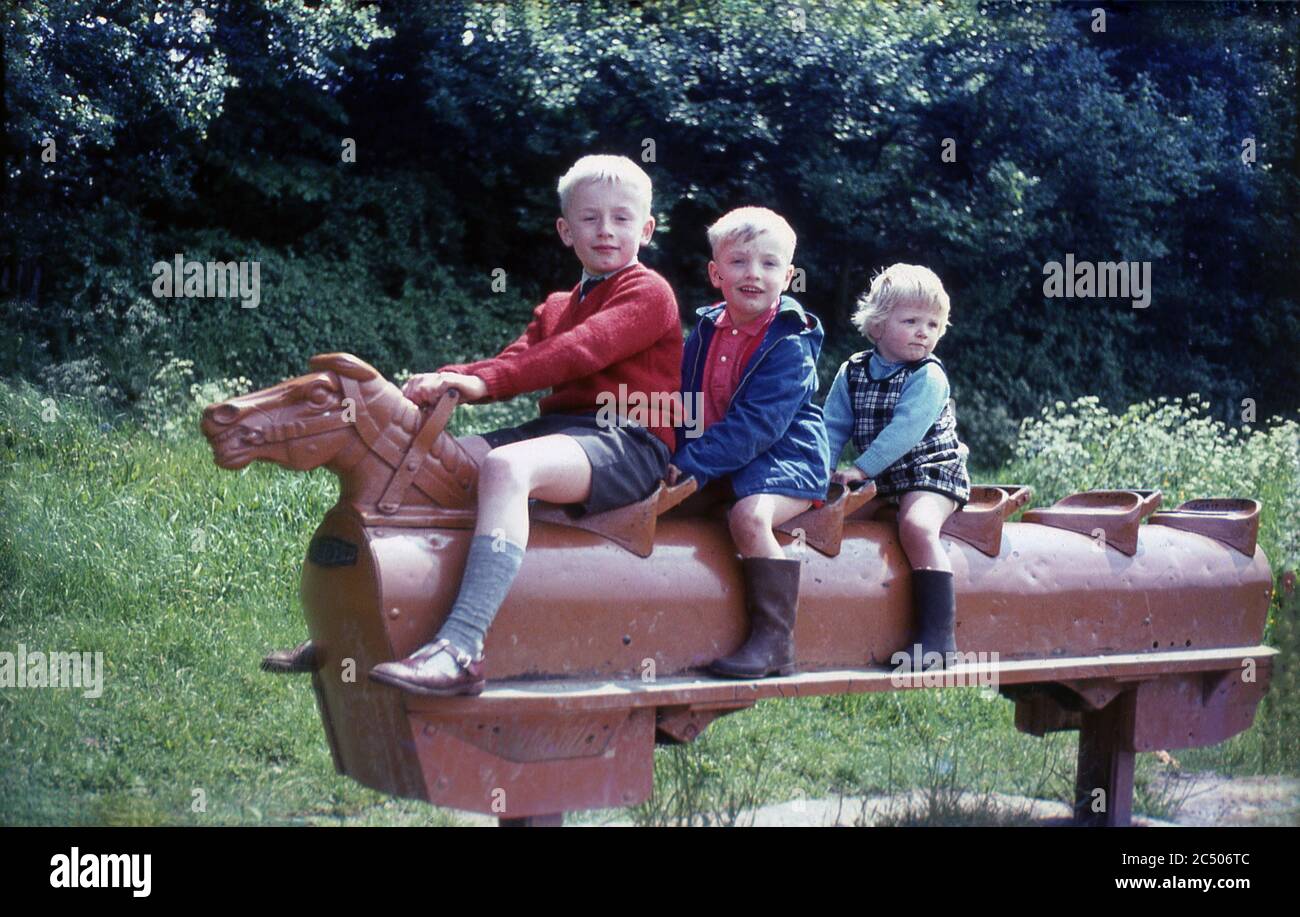 1967, three young children playing or riding on a brown coloured metal five seater rocking horse outside in a park, Surrey, England, UK. This type of traditional, possibly victorian, play equipment was to be found in playgrounds and public parks across Britain in this era and young children had enormous fun playing on them. Stock Photo