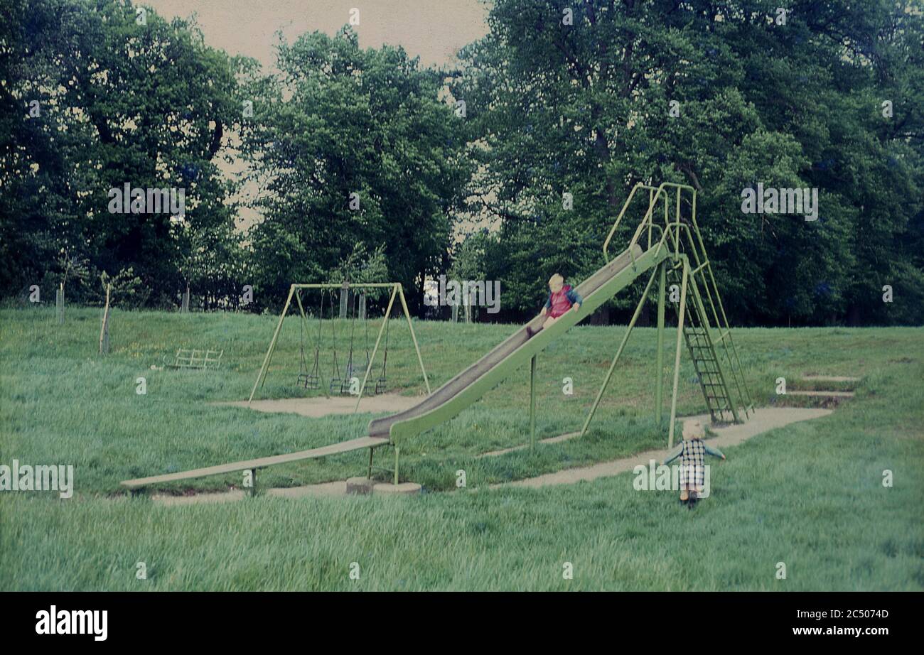 1967, two small children playing outside at a public park or recreation ground, with one child on a traditional metal slide. Old fashioned steel built playground equipment, often dating back to victorian times, was a common sight in parks across Britain in this era, often located as seen here, randomly positioned in grassy areas without too much planning. Stock Photo