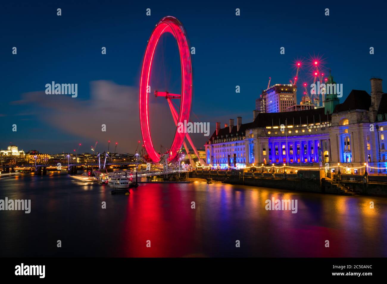 The London Eye and County Hall building at night casting colourful reflections on the Thames river. Stock Photo