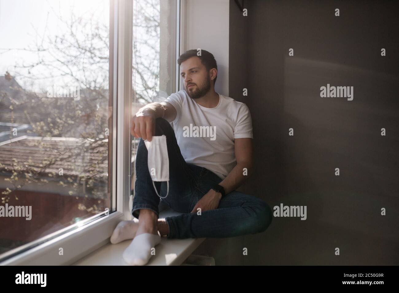 Young man sits on windowsill and hold in hands protective mask and ...