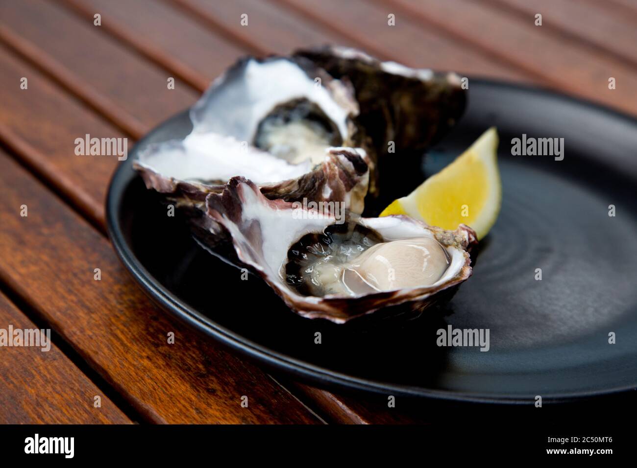 Close up of freshly shucked, chilled Pacific oysters on half shell with lemon wedge on black platter placed on casual wood slats table Stock Photo