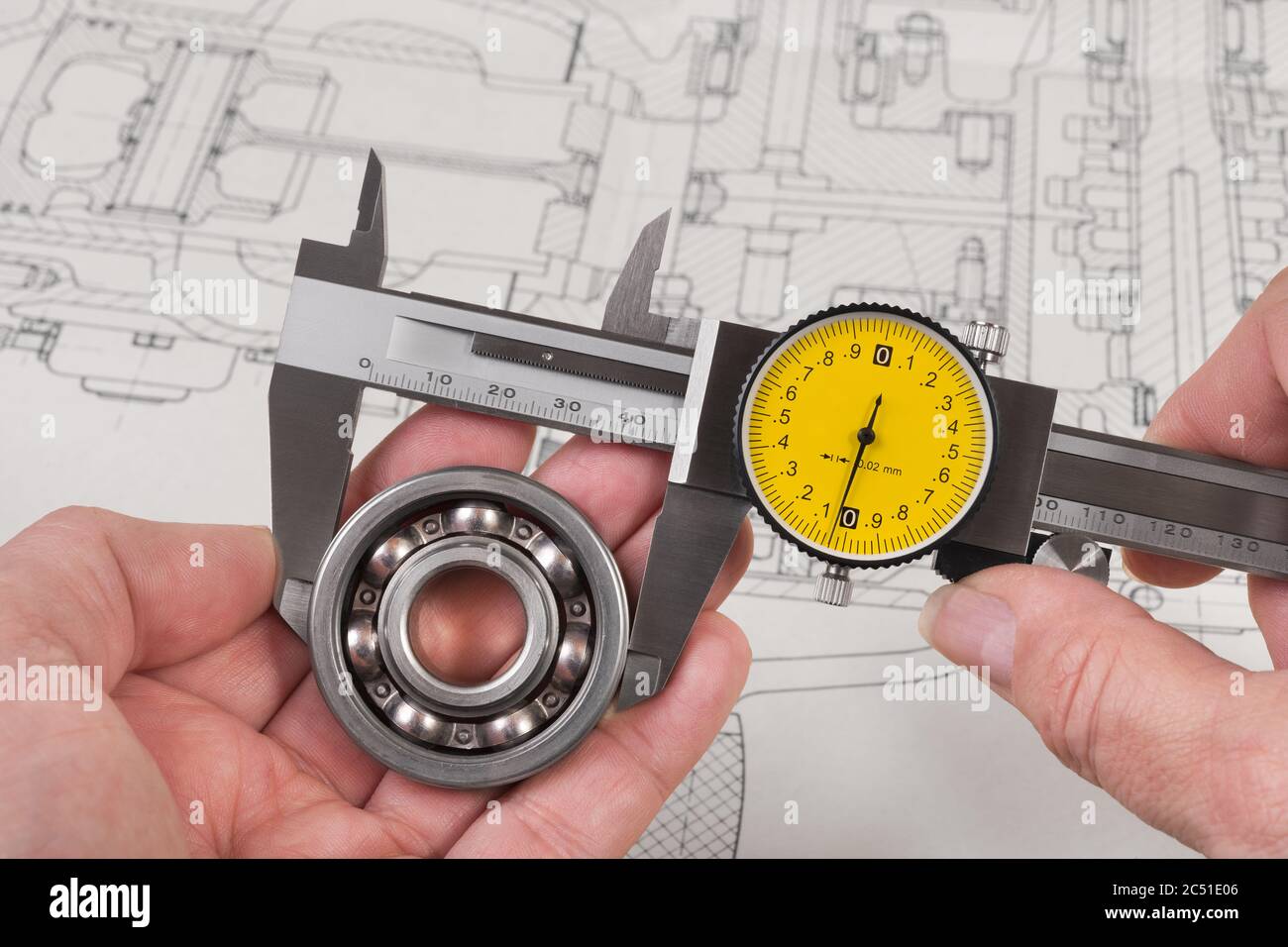 Measurement of steel ball bearing by precise analog caliper above technical drawing. Metallic measuring tool with round yellow dial in engineer hands. Stock Photo