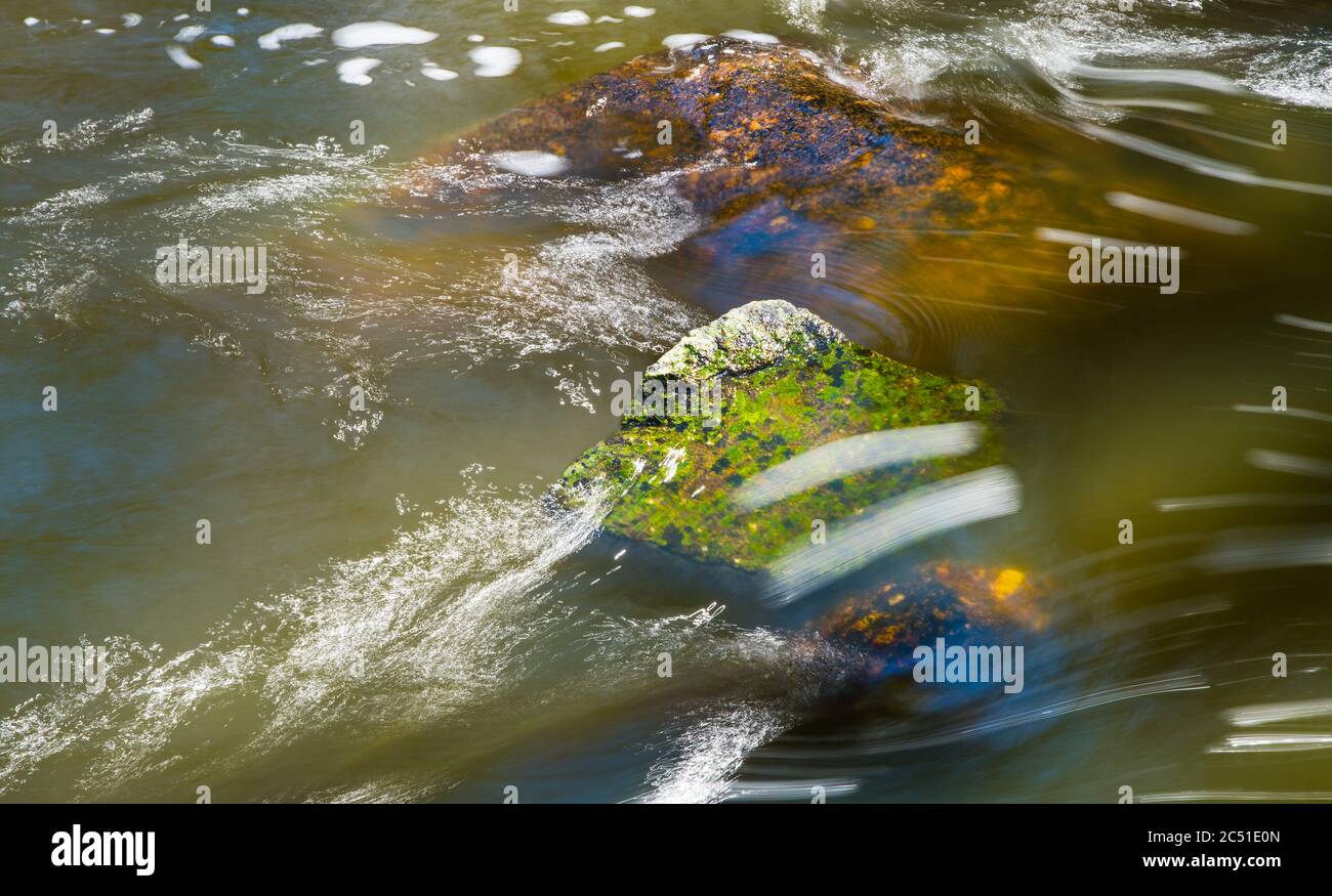 Abstract stream of muddy water flowing over green and brown boulder in wild river bed. Close-up of spring thaw. Playful light reflections, wet stones. Stock Photo