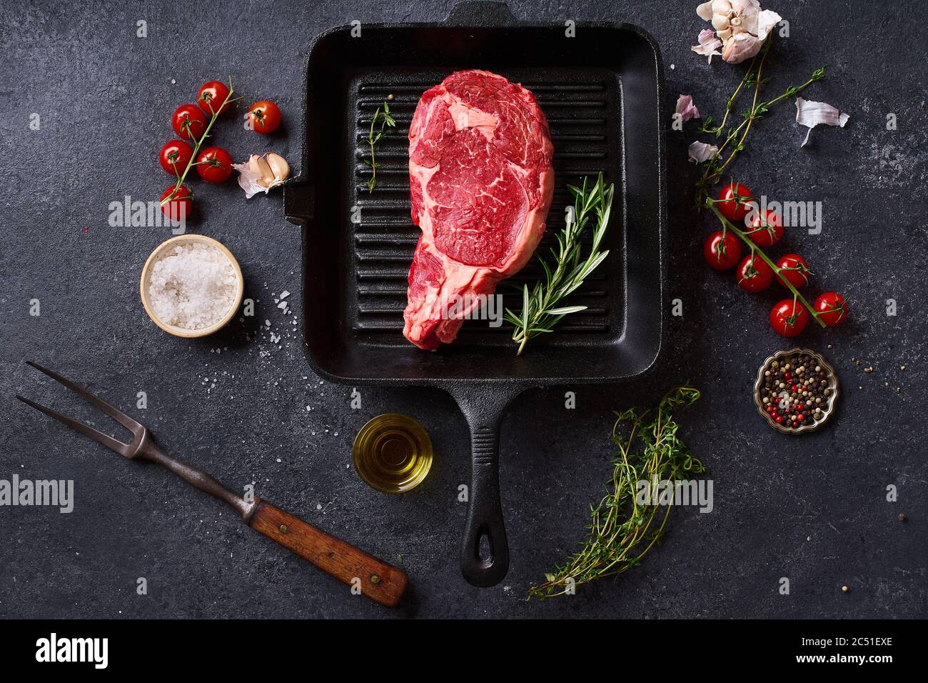 Top view Black Angus prime beef rib eye steak on cast iron grill skillet with fresh rosemary, cherry tomatoes, olive oil and spices. Creative layout w Stock Photo