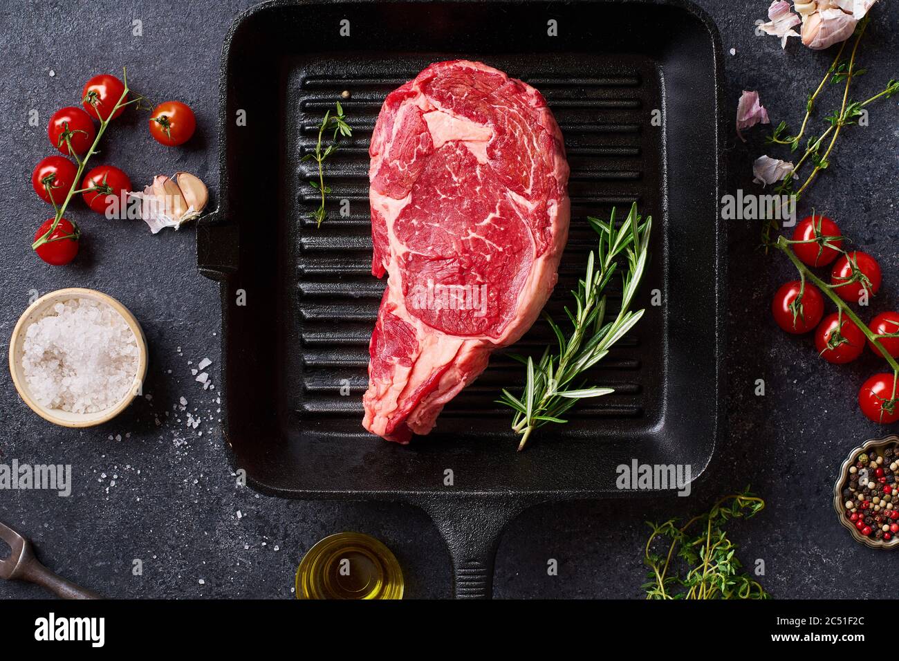 Top view Black Angus prime beef rib eye steak on cast iron grill skillet with fresh rosemary, cherry tomatoes, olive oil and spices. Creative layout w Stock Photo