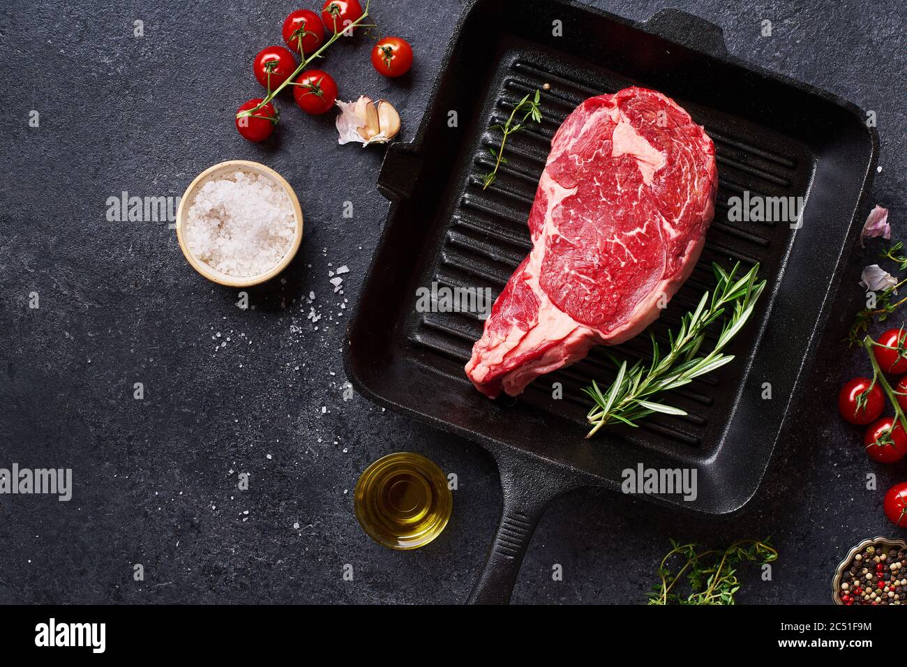 Top view Black Angus prime beef rib eye steak on cast iron grill pan with fresh rosemary, cherry tomatoes, olive oil and spices. Creative layout with Stock Photo