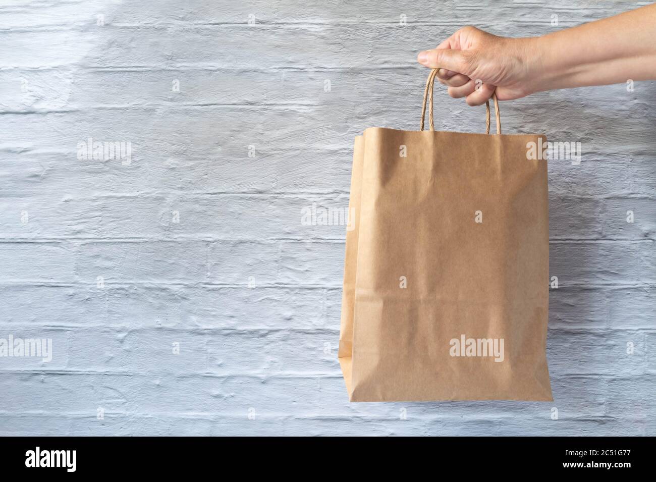 Hand holding paper bag on brick wall background. Paperboard bag in hand on vintage white brickwork wall. Makeup for donation or sponsorship concept Stock Photo
