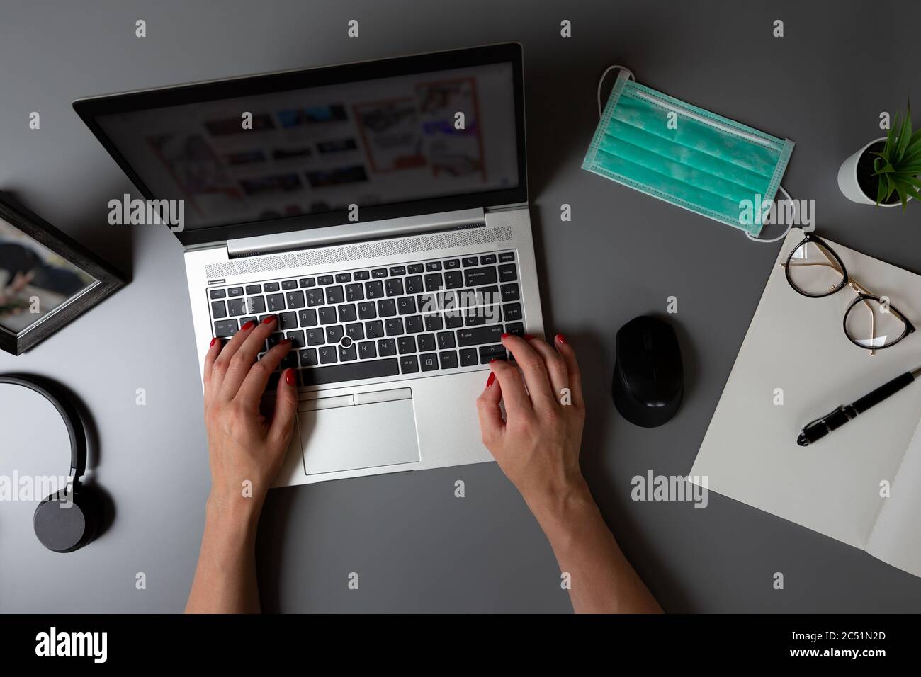 Top view of home office. Women's hands typing on laptop on a gray table with facial protection mask. Stock Photo