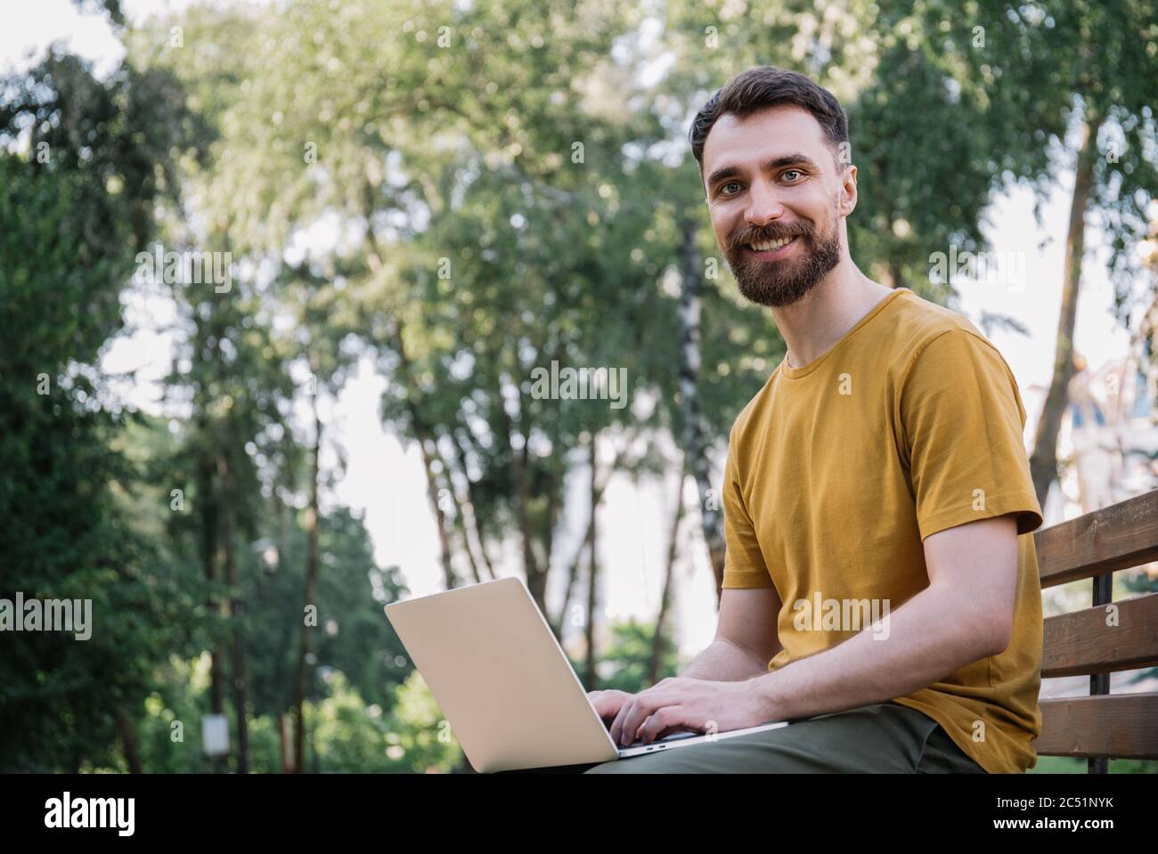 Bearded man using laptop computer, shopping online, sitting on bench. Successful freelancer working in park, typing on keyboard, looking at camera Stock Photo